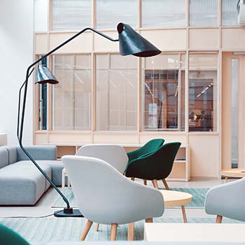 Egy elegáns, minimalista stílusban berendezett világos iroda fehér és zöld székekkel, állólámpákkal és egy fából, meg üvegből készített hátsó fallal a háttérben.